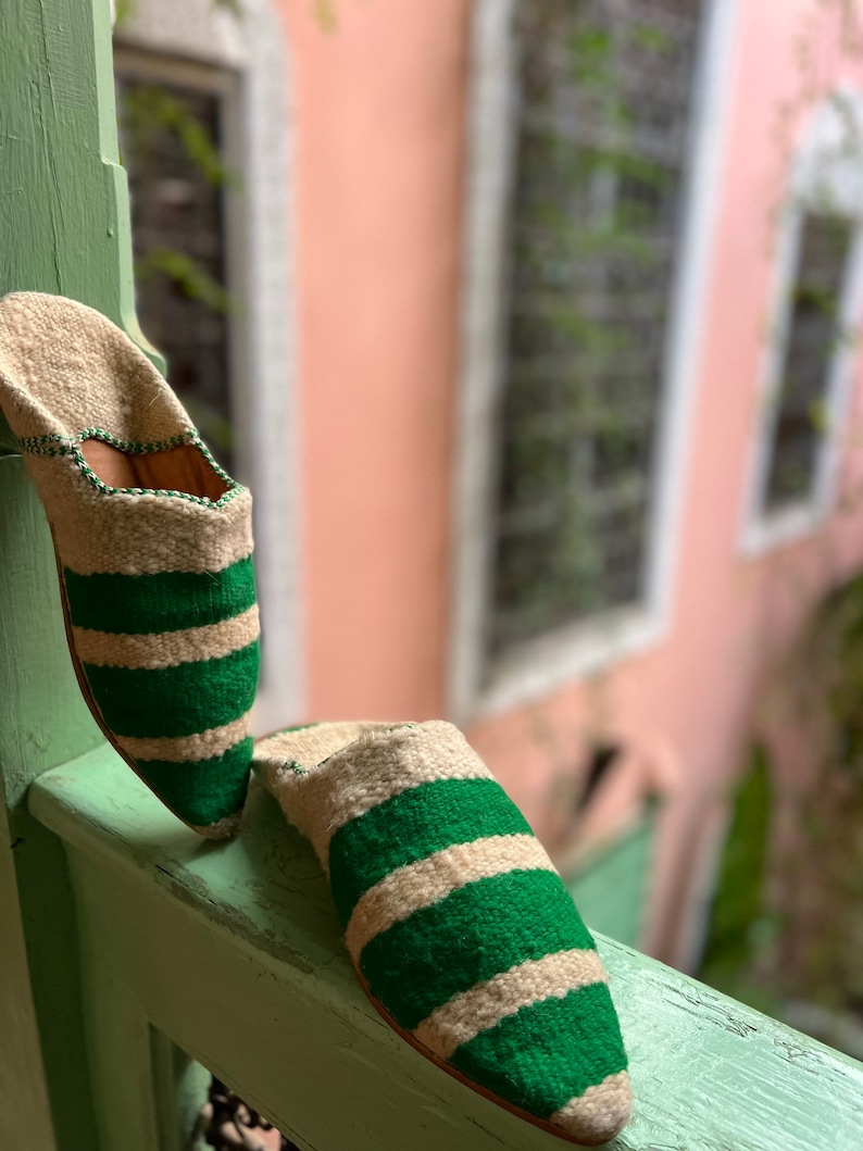 May include: A pair of green and white striped slippers with pointed toes. The slippers are made of a soft, woven fabric and are resting on a wooden surface.