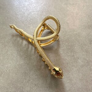 Pu&ograve; includere: Fermaglio per capelli a forma di serpente dorato con finitura testurizzata. Il fermaglio &egrave; a forma di serpente con la coda arricciata e la testa con un muso appuntito.