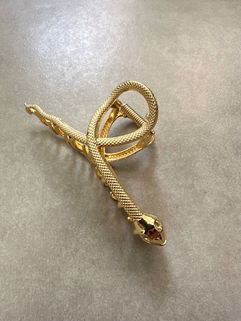 Pu&ograve; includere: Fermaglio per capelli a forma di serpente dorato con finitura testurizzata. Il fermaglio &egrave; a forma di serpente con la coda arricciata e la testa con un muso appuntito.