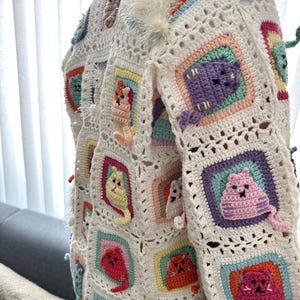 May include: A white crocheted cardigan with colorful cat appliques. The cardigan is made of a soft, lightweight yarn and features a variety of cat designs, including a white cat with a fluffy tail, a purple cat, a yellow cat, and a pink cat.