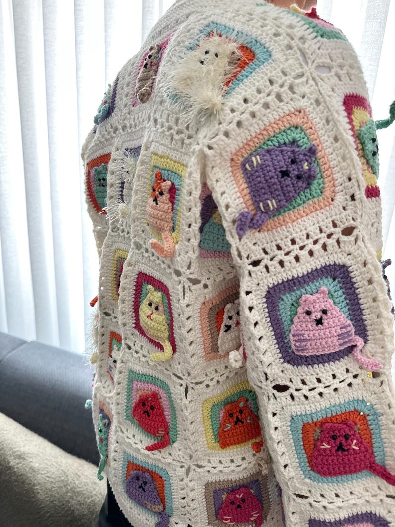 May include: A white crocheted cardigan with colorful cat appliques. The cardigan is made of a soft, lightweight yarn and features a variety of cat designs, including a white cat with a fluffy tail, a purple cat, a yellow cat, and a pink cat.