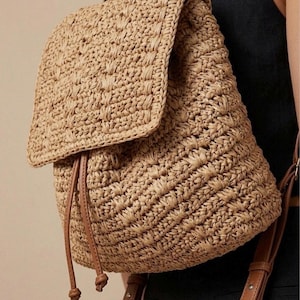 May include: A woven straw backpack in a tan hue, featuring a flap closure and drawstring detail. It has brown leather straps and ties. The bag's design incorporates a textured weave pattern, offering a natural aesthetic.