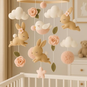 Pode incluir: Móvel de berço com coelhos bege, nuvens brancas, flores cor-de-rosa e um pássaro suspensos por um anel de madeira. O móvel inclui pompons cor-de-rosa e uma estrela, criando uma decoração fantasiosa.