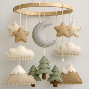 Può includere: Mobile per bambini fatto a mano con stelle, nuvole, una luna crescente, abeti e montagne in feltro. Il mobile è sospeso da un anello di legno, con colori neutri e dettagli cuciti, creando una decorazione incantevole.