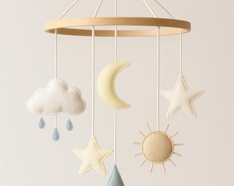 Puede incluir: Móvil para bebé con un aro de madera y formas de fieltro colgantes. Las formas incluyen una nube blanca con gotas de lluvia azules, una luna creciente amarilla, estrellas, un sol y una lágrima azul. El móvil está suspendido por cuerdas blancas.