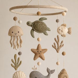 Puede incluir: Móvil para bebé de fieltro con criaturas marinas. Incluye una medusa, tortuga, pez, estrella de mar, ballena, concha y coral. Las criaturas son en tonos neutros de beige, verde y gris, suspendidas de un marco circular con cuentas de madera.