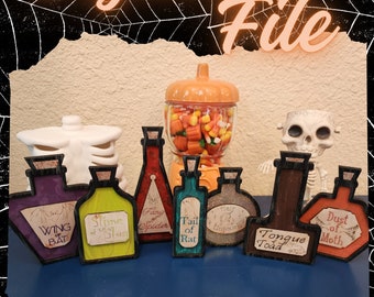 May include: Eight small, wooden potion bottles with Halloween-themed labels. The labels include "Wing of Bat", "Slime of Slug", "Fang of Spider", "Tail of Rat", "Hair of Unicorn", "Tongue of Toad", and "Dust of Moth".