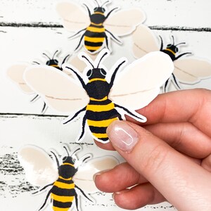 May include: A collection of bee-shaped stickers. Each sticker features a black and yellow striped body, black head, and cream-colored wings. The stickers are on a white wooden surface.