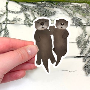 May include: A sticker featuring two cartoon otters. One otter is waving, and the other is looking forward. The otters are brown with light brown faces and white highlights. The sticker is on a white background with decorative greenery.