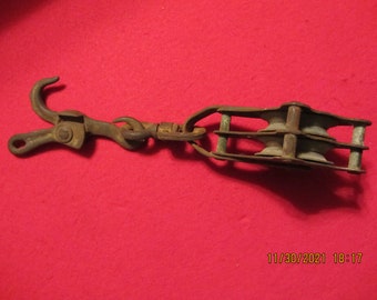 May include: A rusty metal pulley with two wheels and a hook on one end. The pulley is attached to a chain.