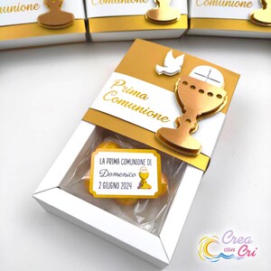 Può includere: Una scatola bianca con un coperchio dorato e un design a calice dorato. La scatola contiene due caramelle gialle e un'etichetta bianca con il testo "LA PRIMA COMUNIONE DI Domenico 2 GIUGNO 2024".