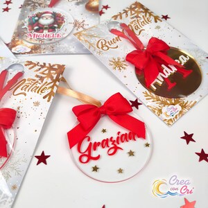 Könnte beinhalten: Weihnachtsschmuck mit roten Schleifen und personalisierten Namen. Ein Ornament zeigt eine Santa Claus-Grafik. Die Ornamente sind transparent mit goldenen Sternakzenten und werden mit dekorativen Karten verpackt.