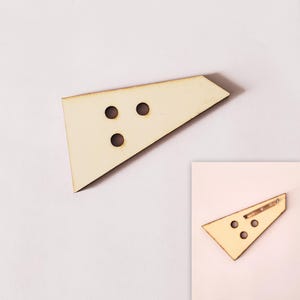 Op de afbeelding: Een lichtgekleurde houten broche in een driehoekige vorm met drie cirkelvormige gaten. De broche heeft een speld op de achterkant voor eenvoudige bevestiging. Het ontwerp is eenvoudig en modern, geschikt voor diverse outfits.