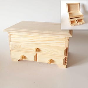 Pode incluir: Uma caixa de madeira com três gavetas e uma tampa com dobradiças. A caixa é feita de madeira clara e tem um design simples. As gavetas têm pequenas maçanetas de madeira.