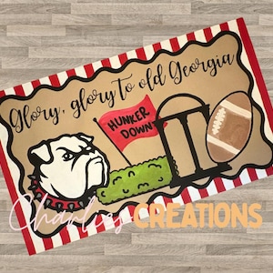 May include: A rectangular sign with a red and white striped border and a brown background. The sign features a bulldog, a football, and the text "Glory, glory to old Georgia." A red flag reads "Hunker Down."