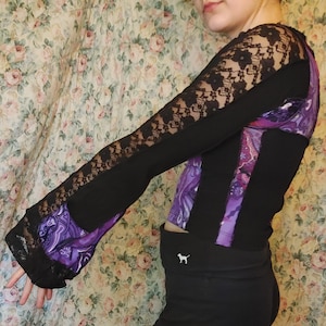 May include: A black long-sleeved shirt with a purple and black paisley print. The shirt has a lace overlay on the sleeves and a black waistband.