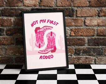 Könnte beinhalten: Gerahmter Druck mit rosa Cowboy-Stiefeln und dem Text "NOT MY FIRST RODEO". Das Design umfasst Kaktus-Silhouetten auf hellrosa Hintergrund. Der schwarze Rahmen kontrastiert mit dem lebendigen Kunstwerk.