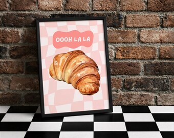 Könnte beinhalten: Ein gerahmter Druck mit einer Aquarellillustration eines Croissants. Der Hintergrund ist ein rosa-weißes Schachbrettmuster mit den Worten "OOOH LA LA" in einer rosa Blase. Der Rahmen ist schwarz.