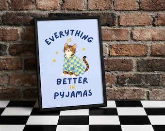 Könnte beinhalten: Gerahmter Kunstdruck mit einer Cartoon-Katze in blau-gelbem Pyjama. Die Katze sitzt auf einem hellblauen Hintergrund mit dem marineblauen Text "EVERYTHING IS BETTER IN PYJAMAS". Goldene Sterne sind ebenfalls im Design enthalten.