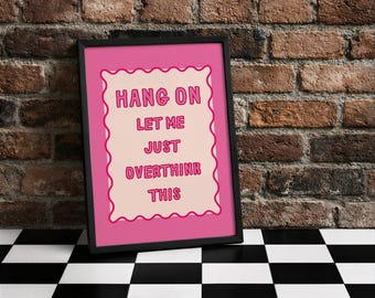 Könnte beinhalten: Ein gerahmter rosa Druck mit den Worten "HANG ON LET ME JUST OVERTHINK THIS" in einer verspielten Schriftart. Der Druck hat einen hellrosa Hintergrund und einen dekorativen Rand. Der Rahmen ist schwarz, und der Druck ist vor einer Backsteinmauer und einem schwarz-weiß karierten Boden platziert.