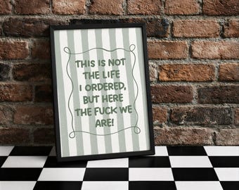 Könnte beinhalten: Ein gerahmter Druck mit dem Text "THIS IS NOT THE LIFE I ORDERED, BUT HERE THE FUCK WE ARE!" in einer verspielten Schriftart. Der Hintergrund des Drucks hat vertikale Streifen. Der Rahmen ist schwarz, und der Druck wird vor einer Backsteinmauer präsentiert.