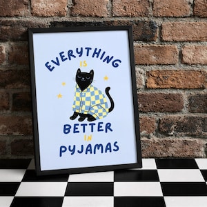 May include: Framed art print featuring a black cat wearing pajamas with a blue and yellow checkered pattern. The text "Everything is better in pyjamas" is written in blue. The background is light blue with yellow star accents.