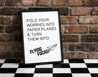 Könnte beinhalten: Ein gerahmter Druck mit dem Text "FOLD YOUR WORRIES INTO PAPER PLANES & TURN THEM INTO FLYING FUCKS!" in schwarzer Schrift. Der Druck zeigt eine Papierflugzeug-Grafik und wird vor einer Backsteinmauer präsentiert.