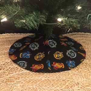 May include: A black Christmas tree skirt with a repeating pattern of the Hogwarts house crests. The crests are in their respective house colors: Gryffindor (red and gold), Hufflepuff (yellow and black), Ravenclaw (blue and bronze), and Slytherin (green and silver).