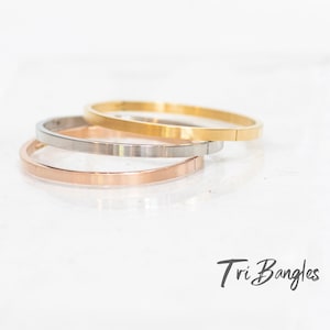 Könnte beinhalten: Drei dünne Metallarmbänder, die übereinander gestapelt sind. Das obere Armband ist golden, das mittlere Armband ist silberfarben und das untere Armband ist roségold.