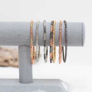 Könnte beinhalten: Ein Set aus acht Perlenarmbändern in Silber-, Gold- und Kupfertönen, die auf einem grauen Ständer präsentiert werden. Die Armbänder bestehen aus kleinen, runden Perlen.