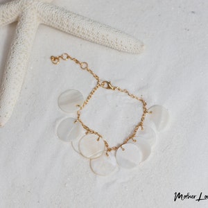 May include: A gold chain bracelet with white mother of pearl round charms. The bracelet is on a white sandy surface.