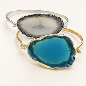 May include: Two bangle bracelets featuring large, irregular agate slices. One bracelet has a silver-tone band with a white and grey agate slice, while the other has a gold-tone band with a vibrant, solid blue agate slice with a metallic edge.