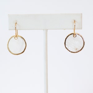 May include: A pair of gold hoop earrings with a white, textured, circular pendant. The earrings are displayed on a white stand.