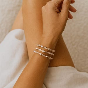 Könnte beinhalten: Ein Set aus drei zierlichen Armbändern am Handgelenk, die jeweils aus einer Reihe kleiner, metallischer, unregelmäßig geformter, flacher, runder und rechteckiger Perlen bestehen, die in einem durchgehenden Muster mit poliertem Glanz aufgereiht sind.