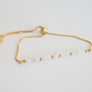 May include: A gold-colored adjustable bracelet featuring a delicate chain and a row of white beads interspersed with small gold-colored beads. The bracelet has a sliding clasp for easy adjustment and a minimalist design.