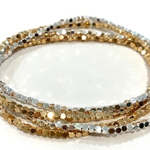 Può includere: Un set di tre braccialetti sovrapposti composti da piccole perline metalliche a forma di cubo. I braccialetti alternano finiture lucide color oro e argento, creando un aspetto stratificato e riflettente con una texture uniforme.
