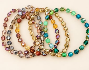 Könnte beinhalten: Eine Sammlung von fünf kreisförmigen Stretch-Armbändern mit facettierten Glasperlen in verschiedenen Farben wie Grün, Blau, Gelb, Braun und Violett, die durchgehend mit kleinen metallisch goldfarbenen Zwischenperlen versehen sind.