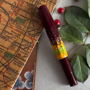 以下が含まれることがあります： 黄緑と白のアクセントがついた花柄の装飾が施された、濃い赤紫色の万年筆。万年筆は、地図と緑の葉を背景にした白い表面の上に置かれている。