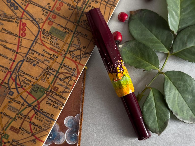以下が含まれることがあります： 黄緑と白のアクセントがついた花柄の装飾が施された、濃い赤紫色の万年筆。万年筆は、地図と緑の葉を背景にした白い表面の上に置かれている。