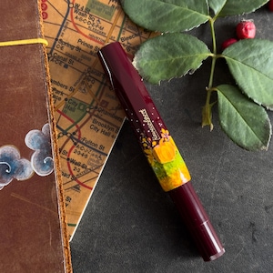 以下が含まれることがあります： 花柄と黄色のデザインが施された、ワインレッドの万年筆。万年筆は、地図と革製のノートが置かれた暗い表面の上に置かれている。