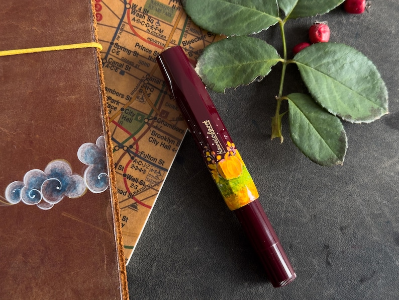 以下が含まれることがあります： 花柄と黄色のデザインが施された、ワインレッドの万年筆。万年筆は、地図と革製のノートが置かれた暗い表面の上に置かれている。