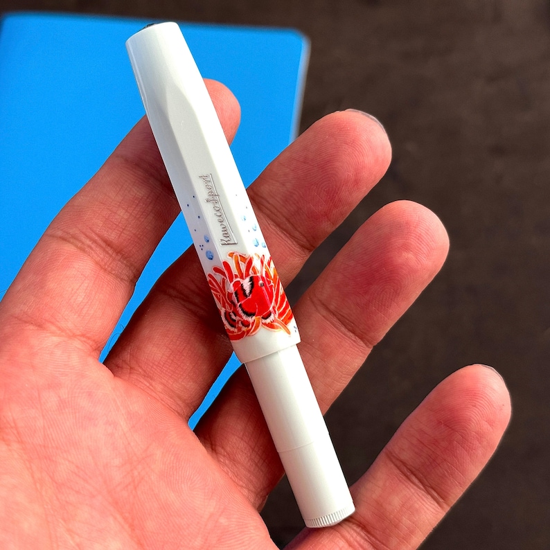 May include: A white pen with a floral design featuring a red flower and blue accents. The pen has the text "Kaweco Sport" printed on the side.