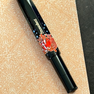 Könnte beinhalten: Ein schwarzer Füllfederhalter mit einem farbenfrohen Design aus Fisch und Koralle. Der Stift trägt die Aufschrift "LAMY" in goldenen Buchstaben.