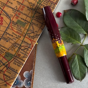 以下が含まれることがあります： 花柄の装飾が施された、ワインレッドとイエローの万年筆。万年筆は、ニューヨーク市の地図の隣にある白い表面の上に置かれています。