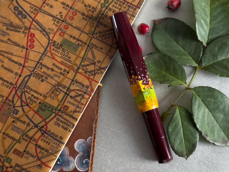 以下が含まれることがあります： 花柄の装飾が施された、ワインレッドとイエローの万年筆。万年筆は、ニューヨーク市の地図の隣にある白い表面の上に置かれています。