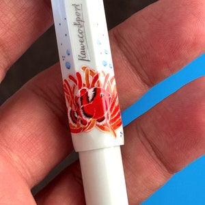 May include: White pen with a red and orange fish design and the text "Kaweco Sport" on the side.