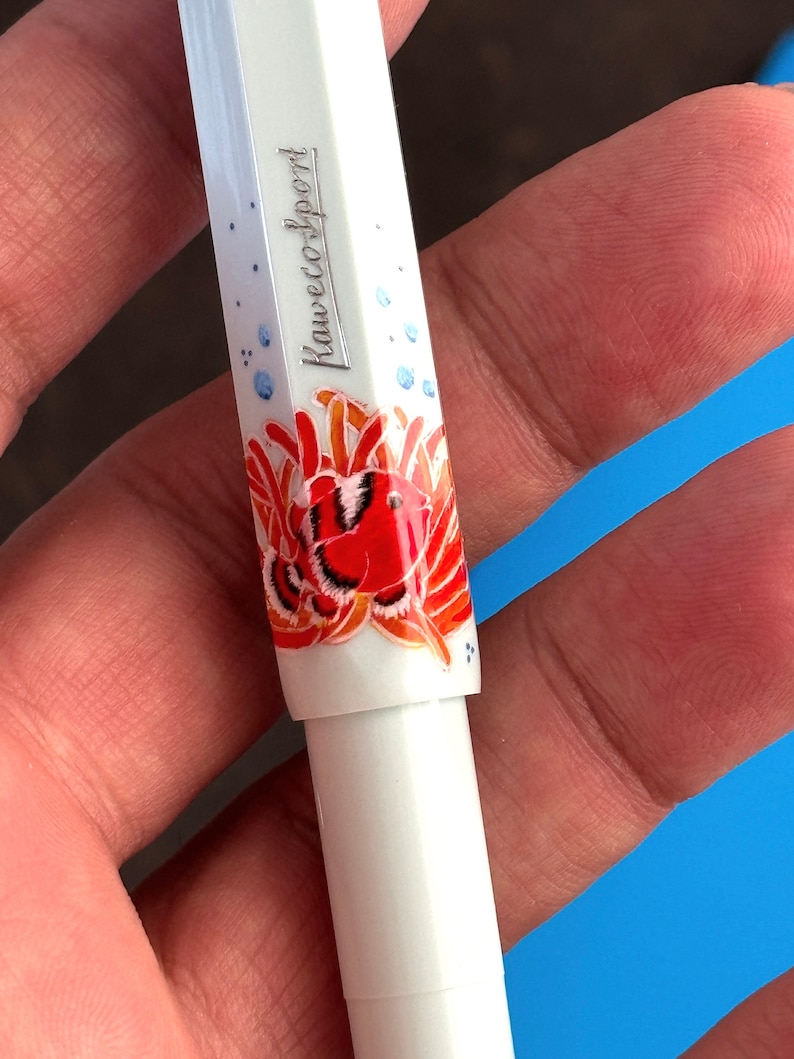 May include: White pen with a red and orange fish design and the text "Kaweco Sport" on the side.