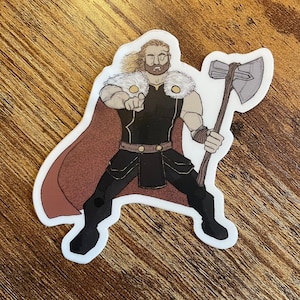 May include: A sticker of a cartoon character with a beard and a cape, holding an axe and pointing at the viewer. The character is wearing a black and gold outfit with a fur collar.