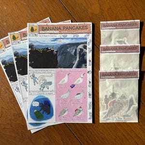 May include: A collection of comic book pages and sticker packs on a wooden surface. The comic book pages feature illustrations and the title "Banana Pancakes." The sticker packs also have the title "Banana Pancakes."