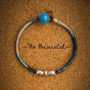 May include: A woven bracelet in shades of blue and white, with a large turquoise bead and silver decorative beads. The text "- Yo Bracelet -" is written in cursive. The bracelet is a stylish accessory.